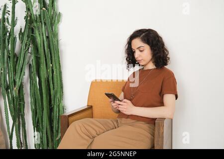Junge hübsche Frau, die sich auf dem Sessel ausruht und online mit ihrem Mobiltelefon spricht Stockfoto
