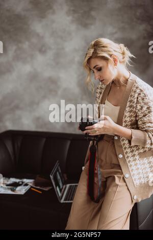 Junge schöne Frau Bloggerin in einem modernen Loft-Interieur. Frau Fotografin Freiberuflerin in beige stilvolle Kleidung schaut auf den Kamerabildschirm.Soft select Stockfoto