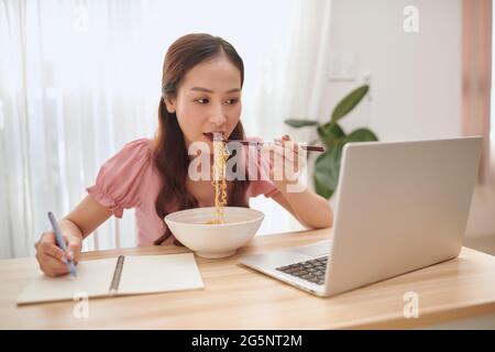 Junge asiatische Frau essen Nudeln und arbeiten mit Laptop zu Hause. Stockfoto