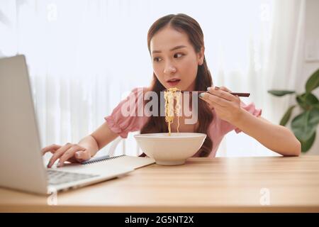 Schöne junge asiatische Frau mit Laptop und asiatische Nudeln zu Hause essen. Stockfoto