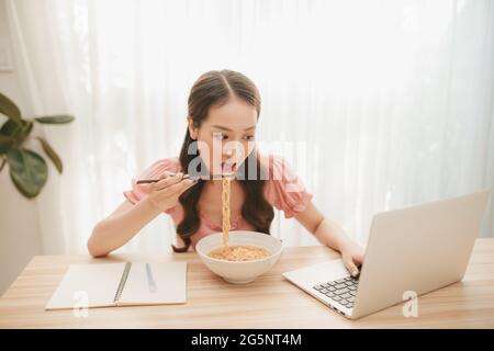 Portrait der jungen asiatischen Frau freiberuflich mit Laptop arbeiten bei Ihr Zuhause und Essen Instant Nudel Stockfoto