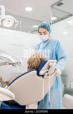 Zahnarzt trägt eine schützende Gesichtsmaske und Einweg-Haarnetz arbeitet mit dem Patienten in der Zahnklinik. Stockfoto