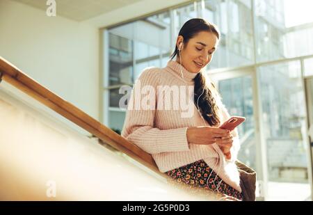 Studentin, die ihr Handy benutzt, während sie an einer Treppe in der Universität steht. Junge Frau, die auf dem College-Campus SMS auf dem Mobiltelefon schreibt. Stockfoto