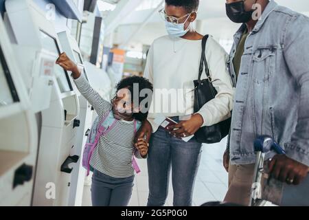 Dreiköpfige Familie am Flughafen in der Nähe des Check-in-Automaten während einer Pandemie. Afrikanisches Paar mit niedlicher Tochter am Flughafen. Stockfoto