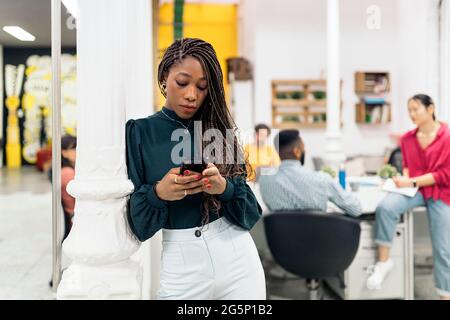 Schwarze Frau mit coolen Zöpfen und formelle Kleidung mit ihrem Handy. Sie arbeitet im Büro. Stockfoto