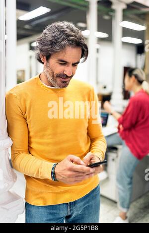 Ein lächelnder Büroangestellter, der sein Mobiltelefon benutzt und im Büro steht. Co-Working-Konzept. Stockfoto