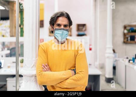 Büroangestellter mit Gesichtsmaske, die seine Arme kreuzt und die Kamera anschaut. Co-Working-Konzept. Stockfoto