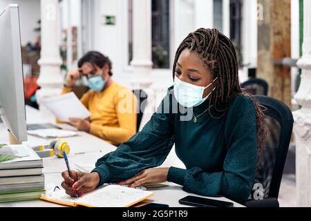 Fokussierte schwarze Frau mit Gesichtsmaske sitzt auf ihrem Schreibtisch und macht Papierarbeit neben anderen Kollegen. Bürokonzept. Stockfoto