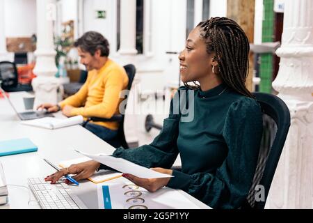 Eine lächelnde schwarze Frau sitzt auf ihrem Schreibtisch und macht neben anderen Kollegen Papierarbeit. Bürokonzept. Stockfoto