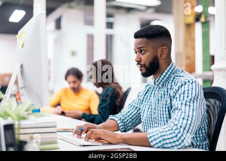 afroamerikanischer Büroangestellter sitzt auf seinem Schreibtisch und benutzt seinen Laptop. Er ist konzentriert und schaut auf die Leinwand. Stockfoto