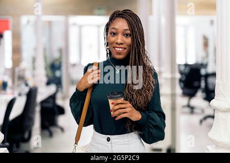 Schwarze Frau mit coolen Zöpfen und in formeller Kleidung lächelnd und mit Blick auf die Kamera. Sie arbeitet im Büro. Stockfoto