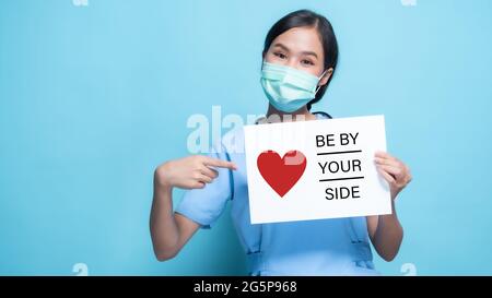 Asiatische, Thai Arzt Frau mit Karte mit Worten AN IHRER SEITE und Stethoskop isoliert auf blauem Hintergrund Stockfoto