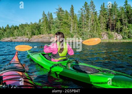 Kajak-Frauen halten Paddel horizontal, während sie eine andere Kajakfahrerin in der Nähe passieren. Sie trägt eine magentafarbene Jacke und eine grüne Sicherheitsweste. Sommerkiefer Stockfoto