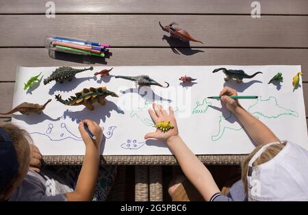 Zwei Kinder zeichnen mit Filzstift kontrastierende Schatten aus Spielzeugdinosauriern. Ideen für die Kreativität der Kinder. Interessante Aktivitäten für Kinder im ho Stockfoto