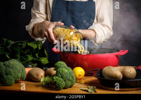 Eine Frau kocht Pasta in einem Topf mit Dampf, auf dem Tisch Brokkoli, Kartoffeln, Kochtopf und Gewürzen Stockfoto