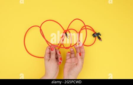 Weibliche Hand hält verdrilltes Kabel zum Laden von mobilen Geräten in einer roten Textilverpackung auf einem gelben Hintergrund, Nahaufnahme Stockfoto