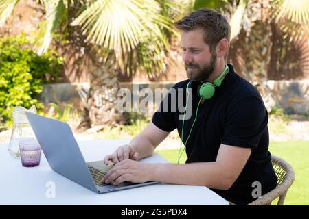 Seitenansicht ein hübscher junger Geschäftsmann, der mit einem Computer aus der Ferne arbeitet und auf der Terrasse am Holztisch sitzt. Stockfoto