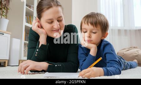 Kleiner Junge und junge Mutter liegen auf dem Boden im Wohnzimmer und machen Hausaufgaben im Copybook. Konzept von Kindererziehung, Entwicklung und glücklicher Elternschaft. Stockfoto