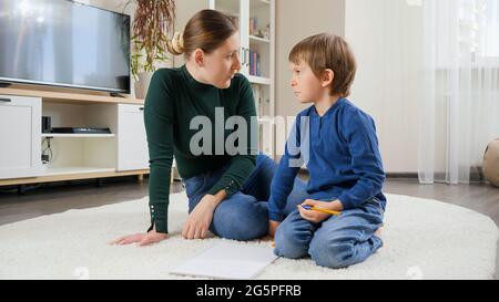 Die junge Mutter ist unzufrieden, schreit und redet streng mit ihrem Sohn, der Hausaufgaben auf dem Teppich im Wohnzimmer macht. Konzept der häuslichen Bildung, Kind Stockfoto