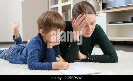 Verärgerte Mutter, die neben dem kleinen Sohn lag, der Hausaufgaben auf dem Teppich machte Stockfoto