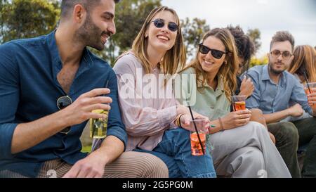 Eine Gruppe von glücklichen besten Freunden, die sich unterhalten, sitzen auf einer Bank im Freien und trinken Cocktails in Plastikgläsern. Junge Leute haben Spaß Konzept. Stockfoto