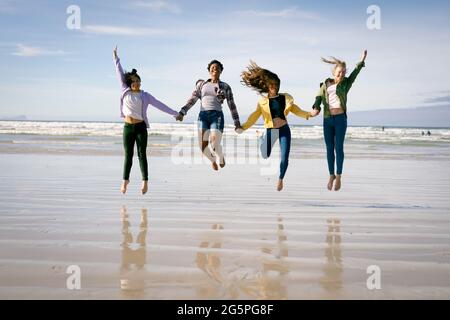 Glückliche Gruppe von verschiedenen Freundinnen, die Spaß haben, am Strand entlang gehen, Hände halten und springen Stockfoto