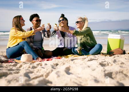 Glückliche Gruppe von verschiedenen Freundinnen, die Spaß haben, am Strand sitzen und Essen lachen Stockfoto