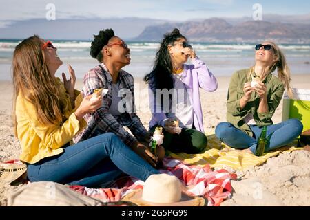 Glückliche Gruppe von verschiedenen Freundinnen, die Spaß haben, am Strand sitzen und essen Stockfoto