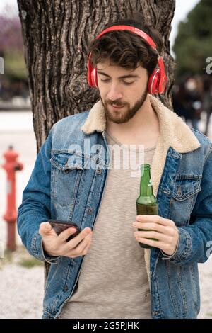 Ein Mann, der Musik mit kabellosen Kopfhörern vom Smartphone hört und ein Bier im Freien trinkt. Stockfoto