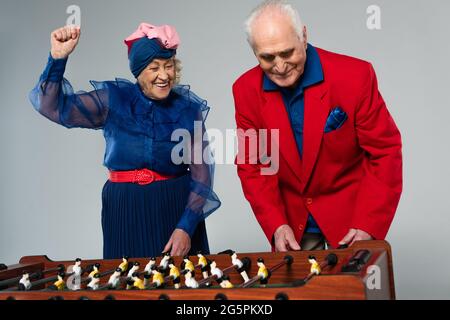 Aufgeregt ältere Paar in stilvoller Kleidung spielen Tischfußball mit Win-Geste auf grau Stockfoto
