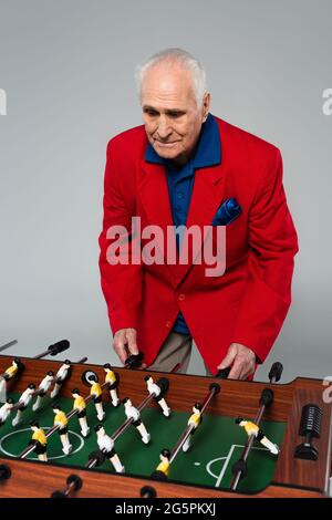 Älterer Mann in rotem Blazer, der Tischfußball auf Grau spielt Stockfoto
