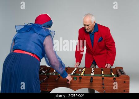 Fröhliches älteres Paar in stilvoller Kleidung, das Tischfußball auf Grau spielt Stockfoto