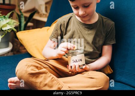Nahaufnahme eines kleinen Jungen, der Hände packt und Stapel-Münzen in ein Glas mit Save-Label legt. Spenden, Geld sparen, Wohltätigkeit, Familie Stockfoto