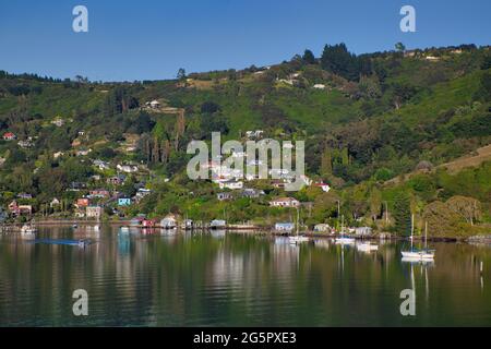 Hübsche Küste mit baumbestandenen Hügeln mit Häusern und Booten direkt vor der Küste, die sich im Meer in der Nähe von Otago Port, South Island, Neuseeland, widerspiegeln Stockfoto