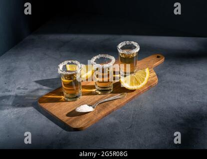 Dunkle Fotografie von Tequila-Aufnahmen mit Salz- und Zitronenscheiben auf einem Holzbrett mit Copyspace. Stockfoto