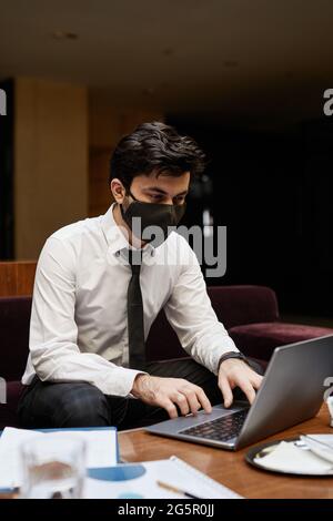 Vertikales Porträt eines jungen Geschäftsmannes, der während der Arbeit in der Hotellobby eine Maske trägt Stockfoto