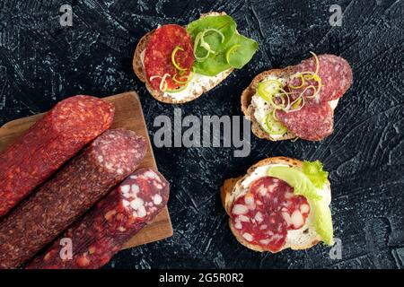 Flach legen drei verschiedene Würste und drei Sandwiches oder Snacks mit ihnen auf einem schwarzen strukturierten Hintergrund. Weichfokus. Stockfoto