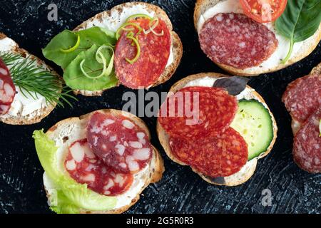 Nahaufnahme von Sandwiches oder Snacks mit verschiedenen Würstchen und Gemüse auf schwarzem Hintergrund. Weicher Fokus. Draufsicht. Flach liegend Stockfoto