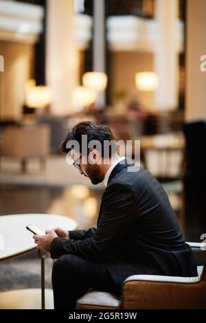 Seitenansicht eines bärtigen Geschäftsmanns aus dem Nahen Osten, der in der Lounge des Hotels auf dem Smartphone wartete Stockfoto