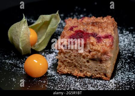 Nahaufnahme eines frisch gebackenen Kuchens mit Himbeeren, Jostaberries und Rhabarber auf einem schwarzen Teller isoliert auf schwarzem Hintergrund mit zwei reifen Physalis Stockfoto