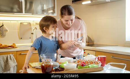 Glücklicher kleiner Junge mit junger Mutter auf der Suche nach einem neuen Rezept zum Kochen auf dem Smartphone. Kinder kochen mit Eltern, kleiner Koch, Familie hat Zeit Stockfoto