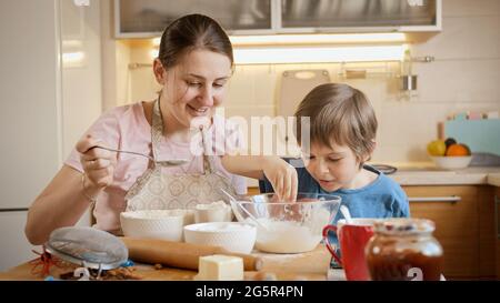 Lächelnder kleiner Junge mit Mutter, die Zutaten in einer großen Schüssel mixt, um Teig oder Keks zu machen. Kinder kochen mit Eltern, kleiner Koch, Familie hat Zeit Stockfoto