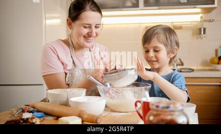 Kleiner lächelnder Junge hält Sieb und hilft der Mutter, Teig für Keks zu machen. Kinder kochen mit Eltern, kleiner Koch, Familie, die Zeit zusammen hat Stockfoto