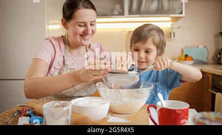 Netter Junge mit Mutter, der Mehl mit Sieb für Kuchen oder Tortenteig sieben. Kinder kochen mit den Eltern, kleiner Koch, Familie, die Zeit zusammen, häusliche Stockfoto