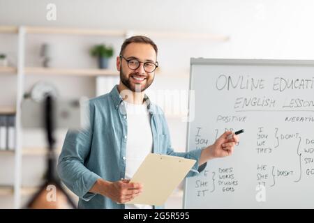 Porträt eines glücklichen jungen Lehrers, der in der Nähe einer Tafel mit englischen Grammatikregeln steht und Internetunterricht durchführt Stockfoto