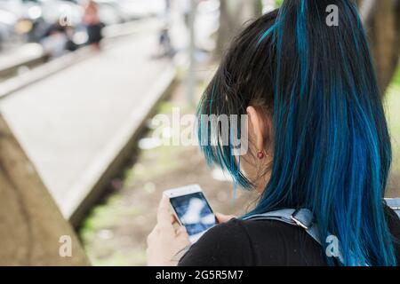 Schöne junge blauhaarige College-Mädchen Blick auf ihre sozialen Netzwerke in einem Park auf einem Baum lehnt. Nahaufnahme des Rückens. Mädchen auf ihr Handy Stockfoto