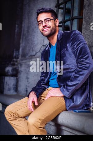Junger Mann, Der Sich Draußen Entspannt. In einem dunkelblauen Wollblazer, einem blauen Pullover und einer dunkelgelben Hose trägt ein junger Mann mit Brot eine Brille Stockfoto