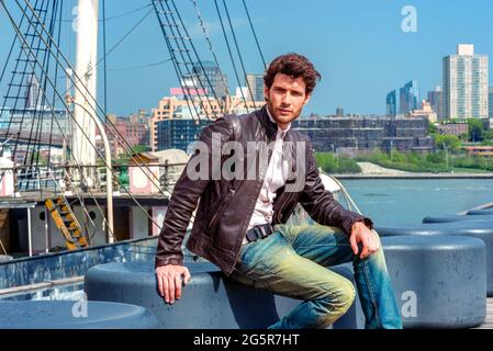 Ein europäischer Geschäftsmann reist in New York City. In einer schwarzen Lederjacke, einer blauen Jeans sitzt ein junger Mann mit Bart auf einer Bank Stockfoto