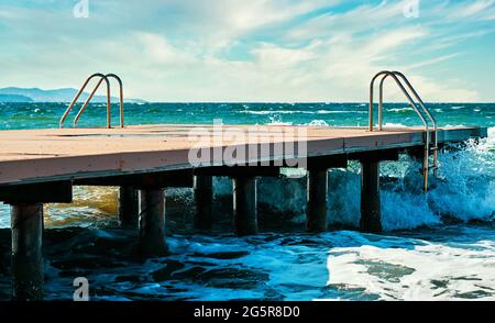 Hölzerner Pier mit Metallleitern, plätschernden Wellen und dem Meer an einem windigen Sommertag. Stockfoto