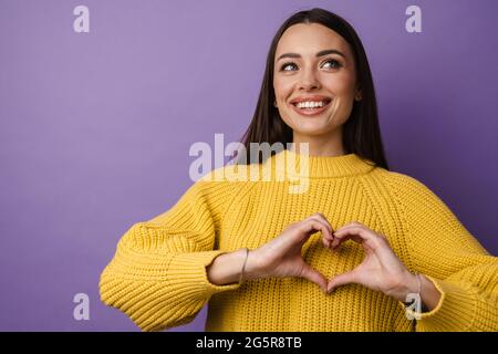 Glückliche junge Frau im Pullover mit Herz-Symbol mit Händen über violettem Wandhintergrund Stockfoto